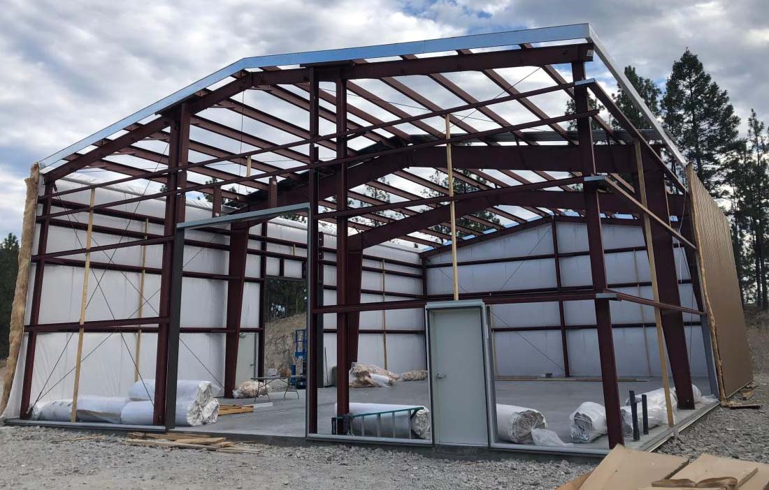Structural steel frame of a prefabricated metal garage under construction, showing red steel beams and wall panels ready for installation.