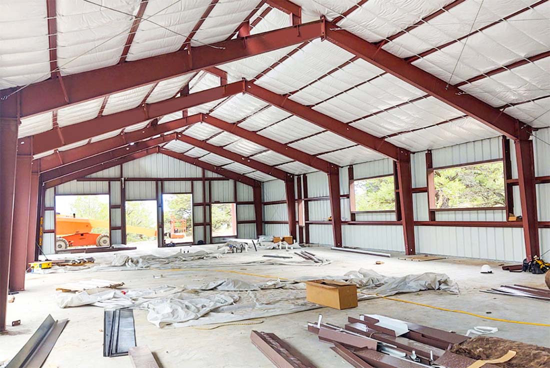 Interior of a clear-span steel barn under construction, showing red steel frames, roof insulation, and wide open space for farm storage and equipment access.