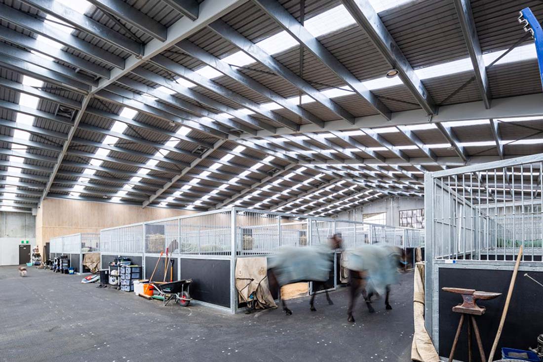Interior view of a steel horse barn with skylight roof panels, metal stall partitions, and wide central aisle for daily stable operations