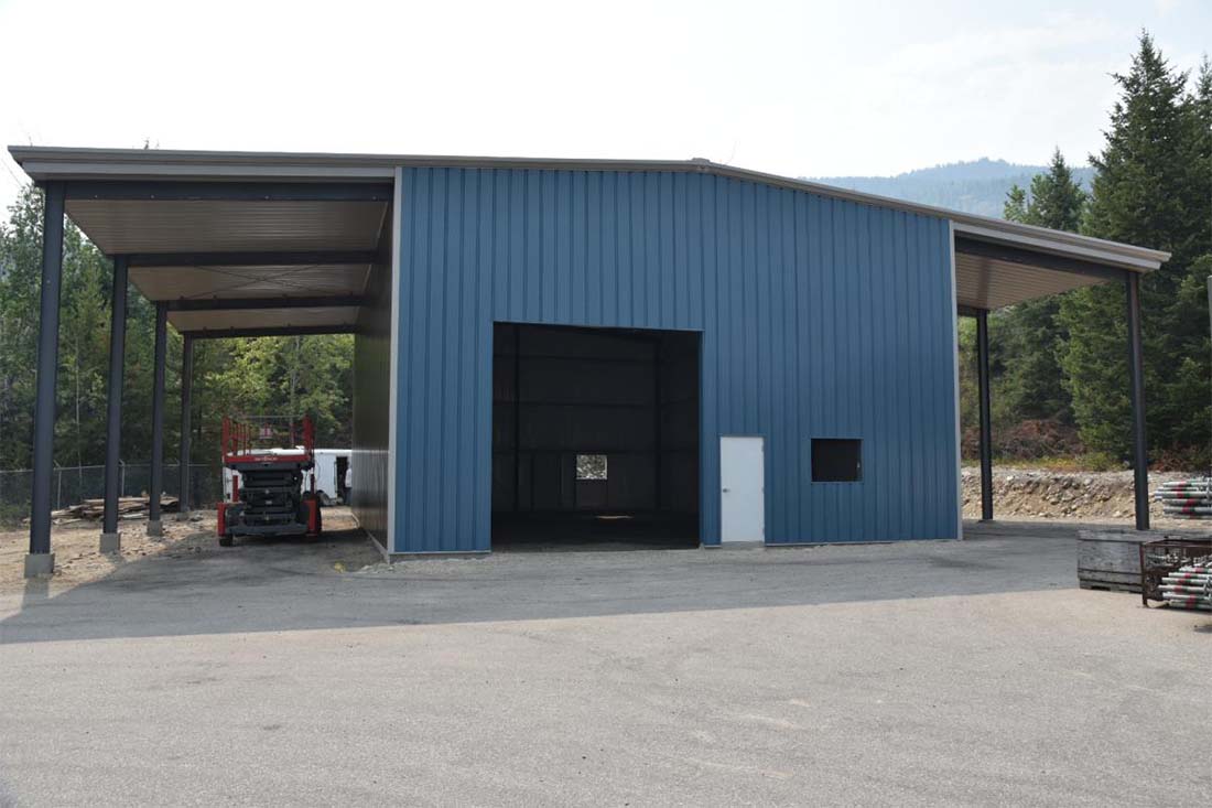 Large metal shed building with blue steel wall panels, lean-to roof extensions on both sides, and a wide front opening for equipment and utility storage.