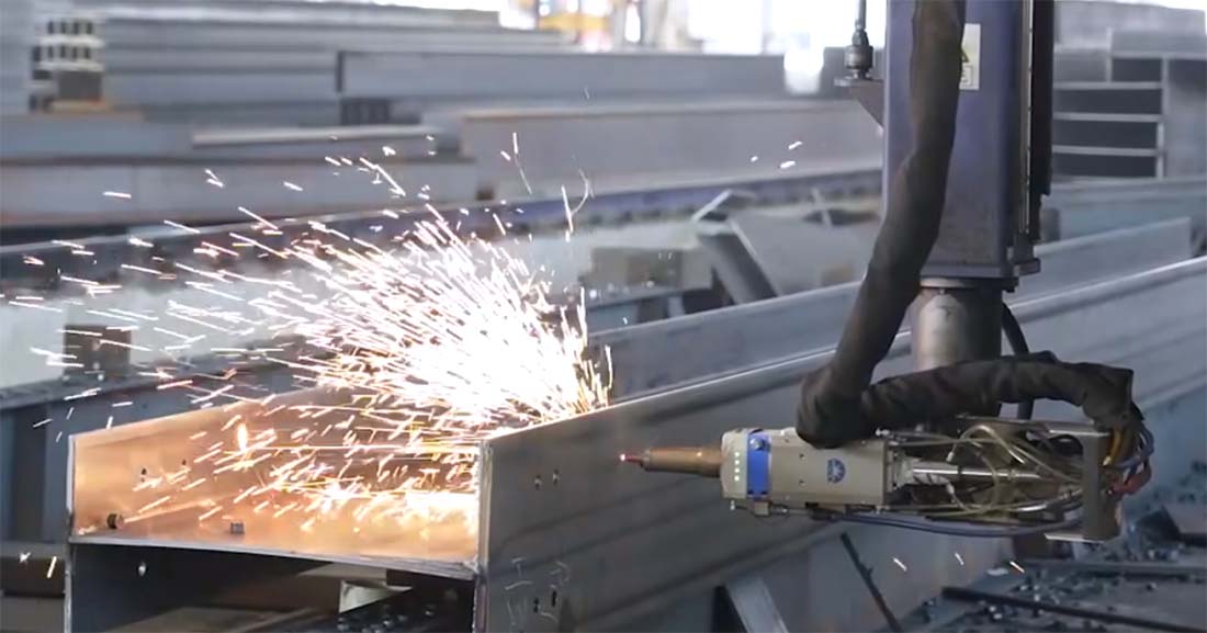 Close-up of precision steel beam fabrication with automated cutting and welding sparks, used to produce structural components for steel barn buildings.