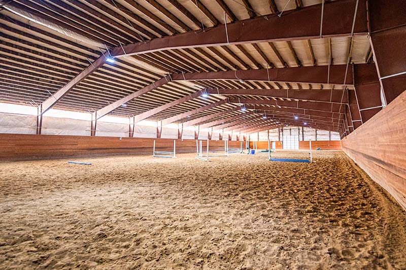 Wide-span metal horse barn arena interior with open space and sand footing for equestrian training.