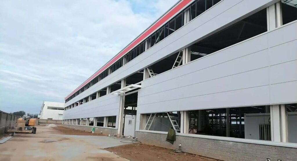 Exterior view of a steel warehouse with wall cladding panels installed, showing enclosure progress, window openings, and trim details.